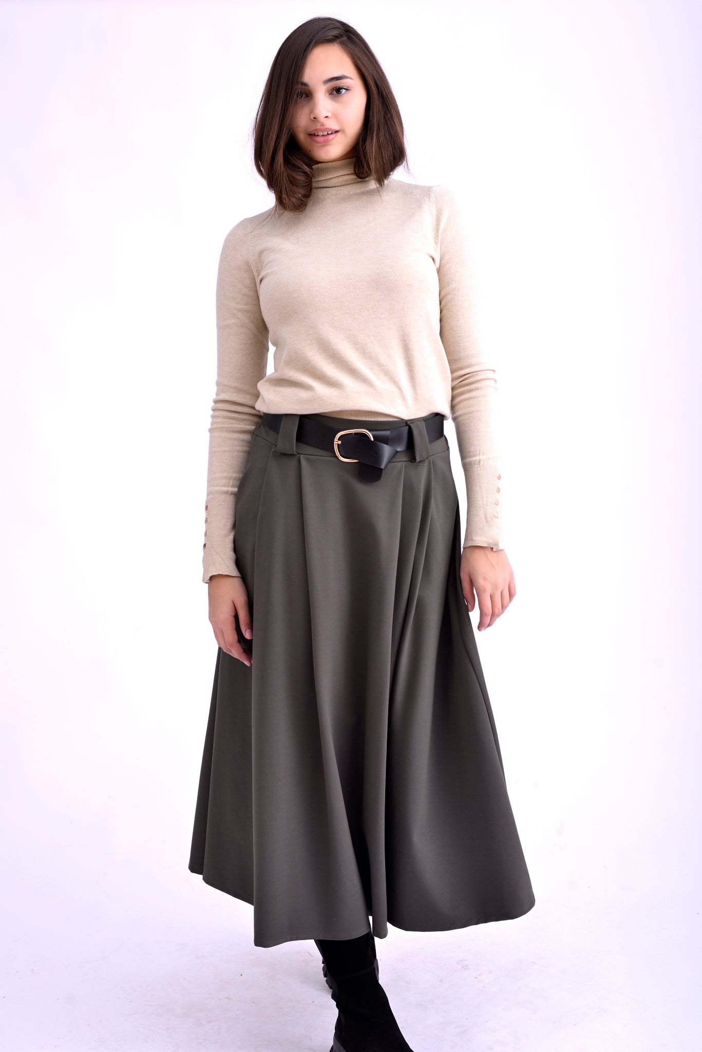 Model purtând o fustă midi gri kaki cu pliuri, curea neagră cu cataramă aurie și pulover bej cu guler înalt, asortată cu ghete negre.