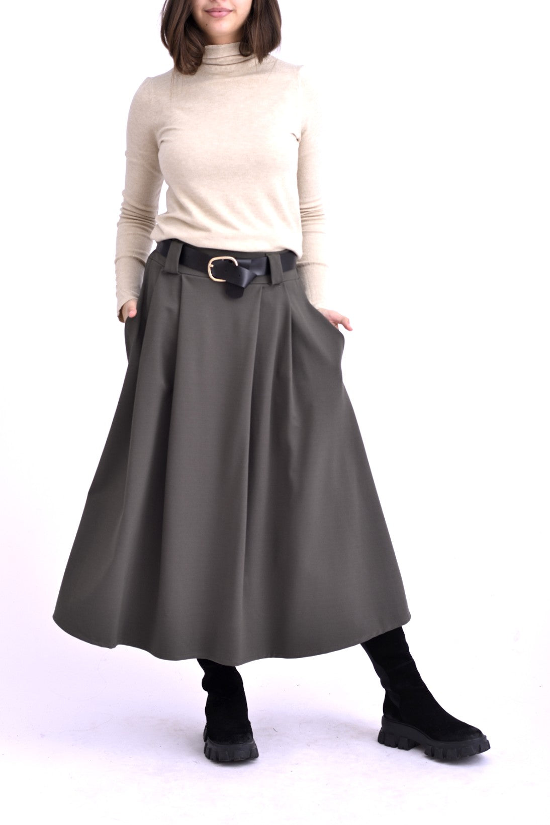Model purtând o fustă midi gri cu pliuri, curea neagră cu cataramă aurie și pulover bej cu guler înalt, asortată cu ghete negre.