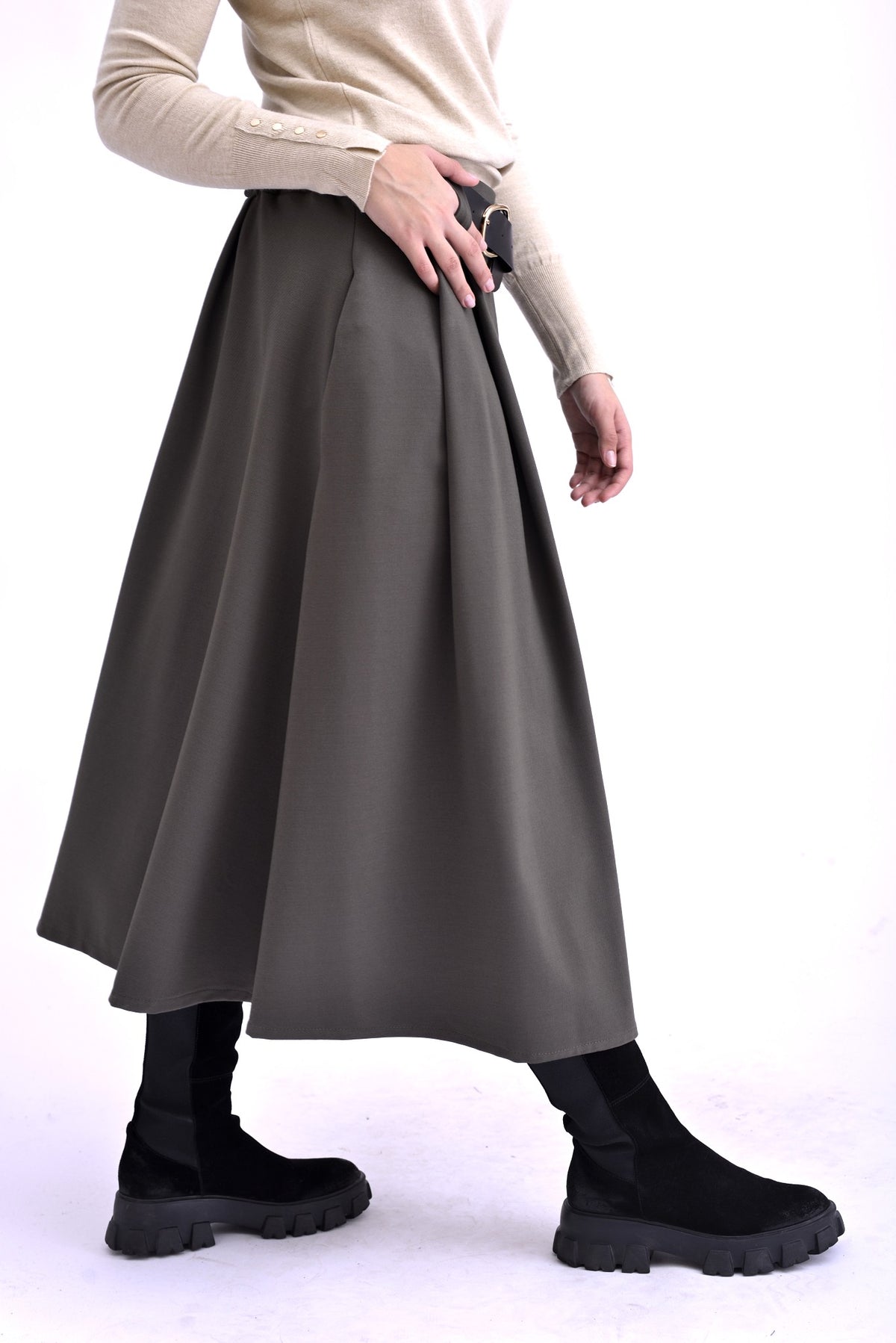 Detaliu lateral al unei fuste gri midi cu pliuri și curea neagră, purtată cu pulover bej și ghete negre.
