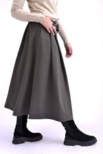 Detaliu lateral al unei fuste gri midi cu pliuri și curea neagră, purtată cu pulover bej și ghete negre.