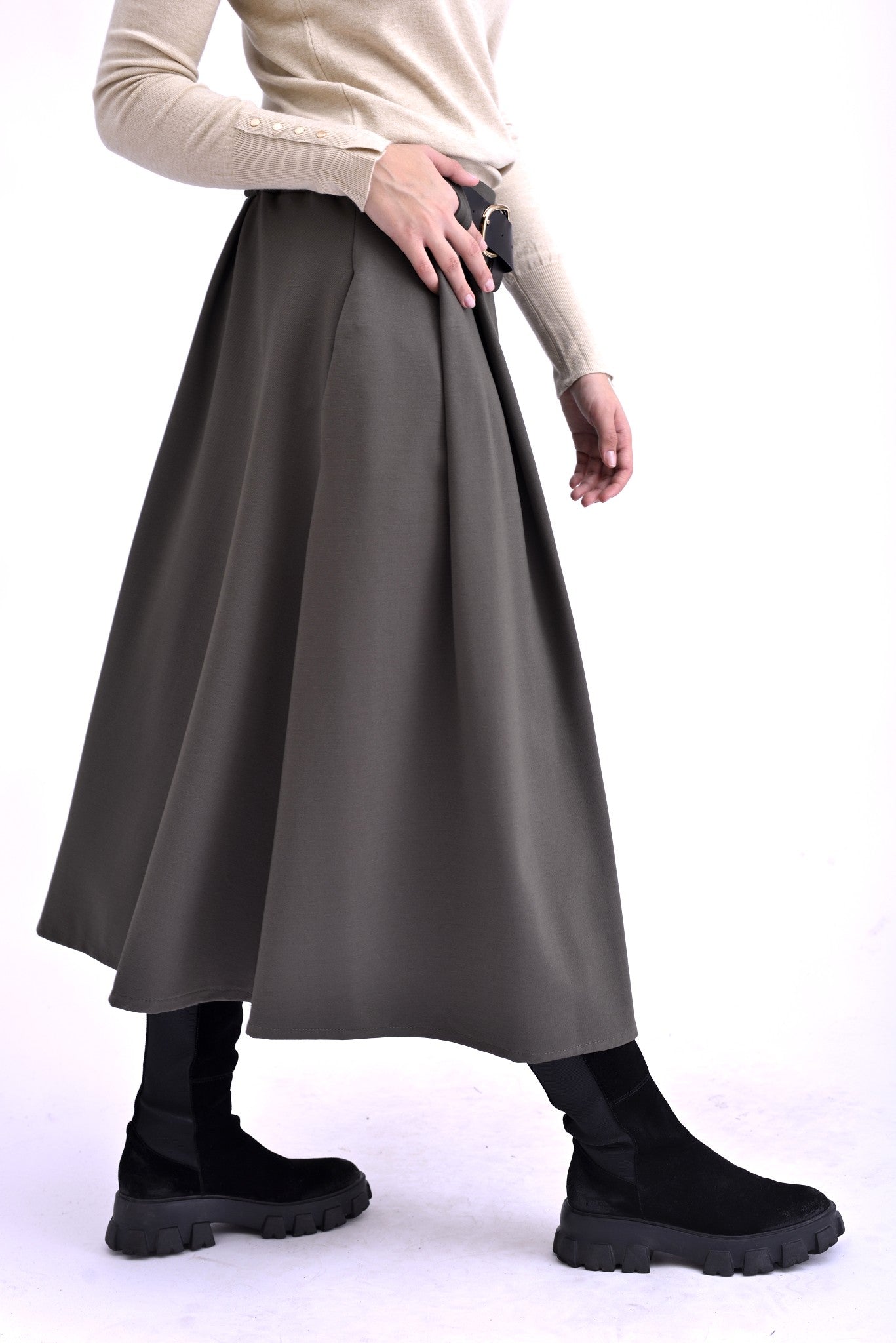 Detaliu lateral al unei fuste gri midi cu pliuri și curea neagră, purtată cu pulover bej și ghete negre.