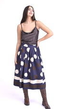 Model purtând o fustă midi maro cu imprimeu cu buline mari în nuanțe de mov și alb, asortată cu un top satinat crem și cizme maro.