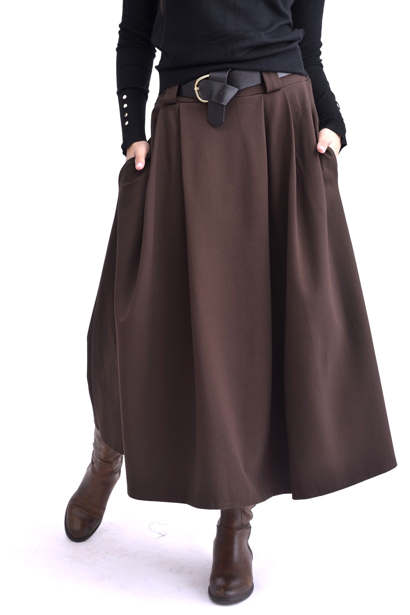 Detaliu al unei fuste maro midi cu pliuri și curea neagră, purtată cu pulover negru și cizme maro.
