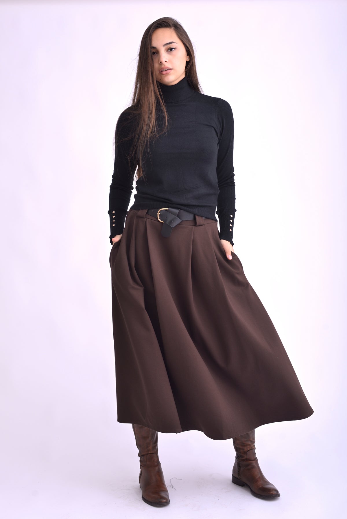 Detaliu al unei fuste maro midi cu pliuri și curea neagră, purtată cu pulover negru și cizme maro.