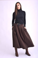 Detaliu al unei fuste maro midi cu pliuri și curea neagră, purtată cu pulover negru și cizme maro.