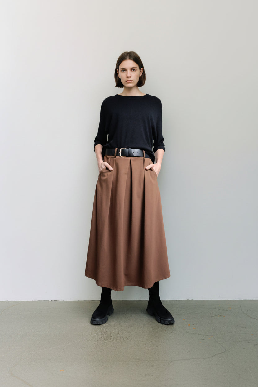 Detaliu al unei fuste maro midi cu pliuri și curea neagră, purtată cu pulover negru și cizme maro.
