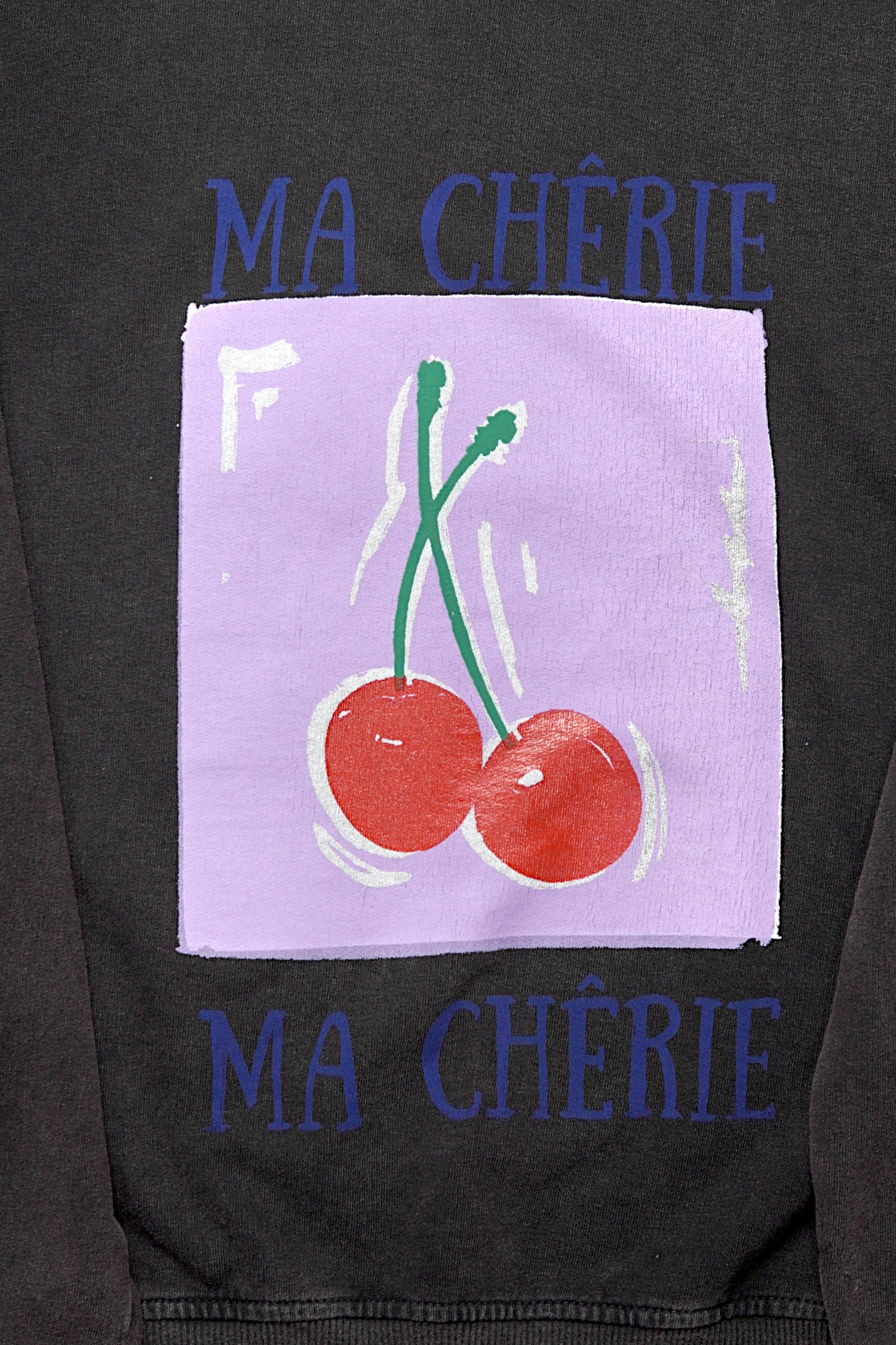 Prim-plan pe imprimeul cu două cireșe roșii și textul „Ma Chérie” de pe hanoracul maro cafea.