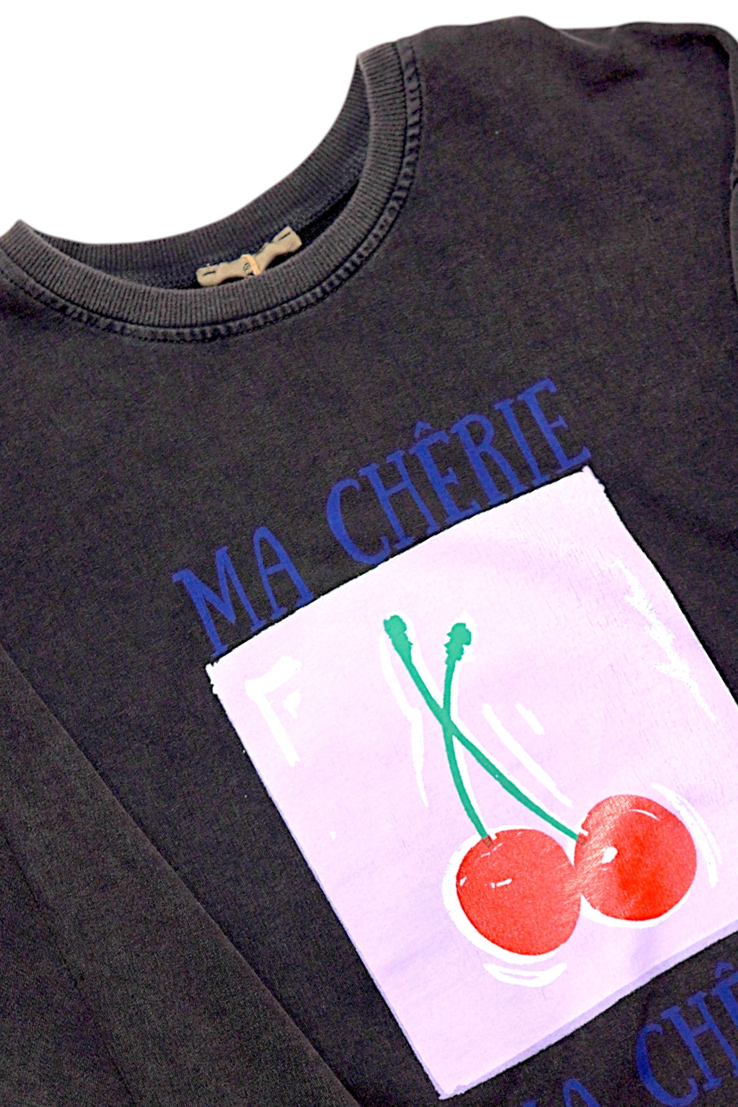 Detaliu de aproape cu gulerul hanoracului maro cafea și imprimeul cu cireșe roșii pe fundal lila și textul „Ma Chérie”.

