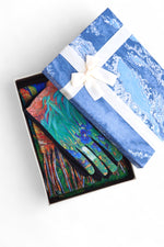 Mănuși colorate cu imprimeu artistic, prezentate într-o cutie cadou albastră cu fundă albă.