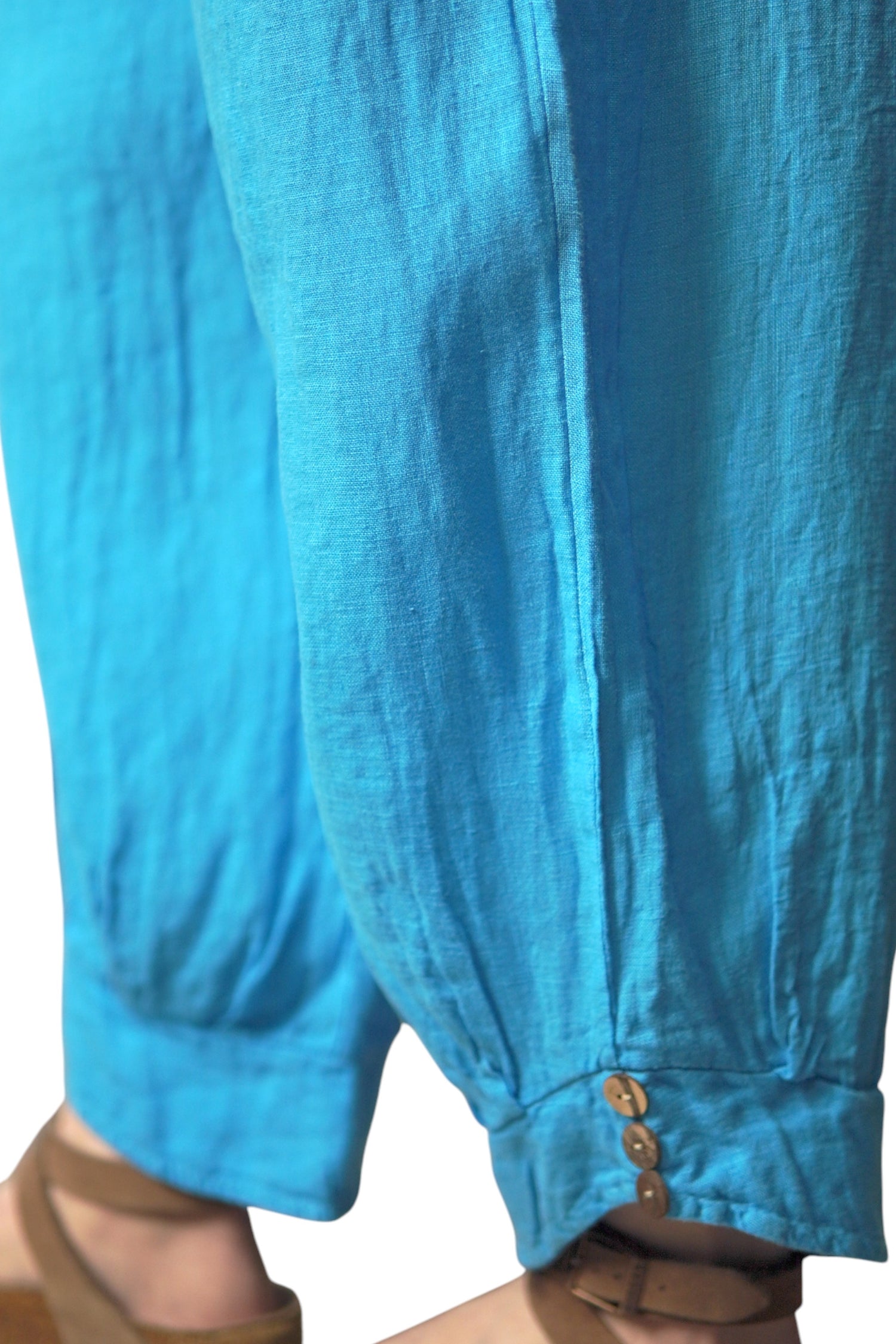 Detaliu al tivului pantalonilor bleu din in, cu încrețituri fine și trei nasturi aplicați.