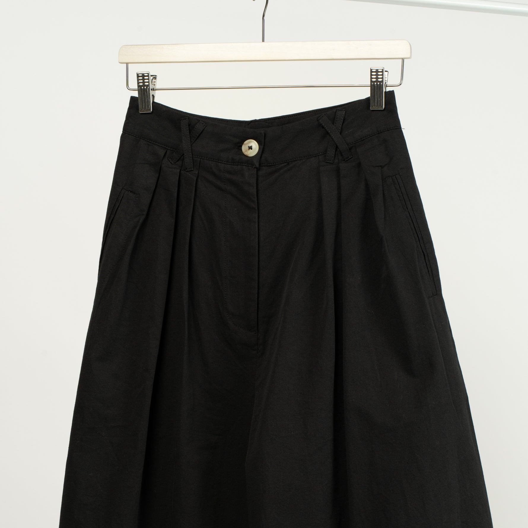 Detaliu al taliei unui pantalon larg negru din bumbac structurat, cu pense și buzunare laterale.