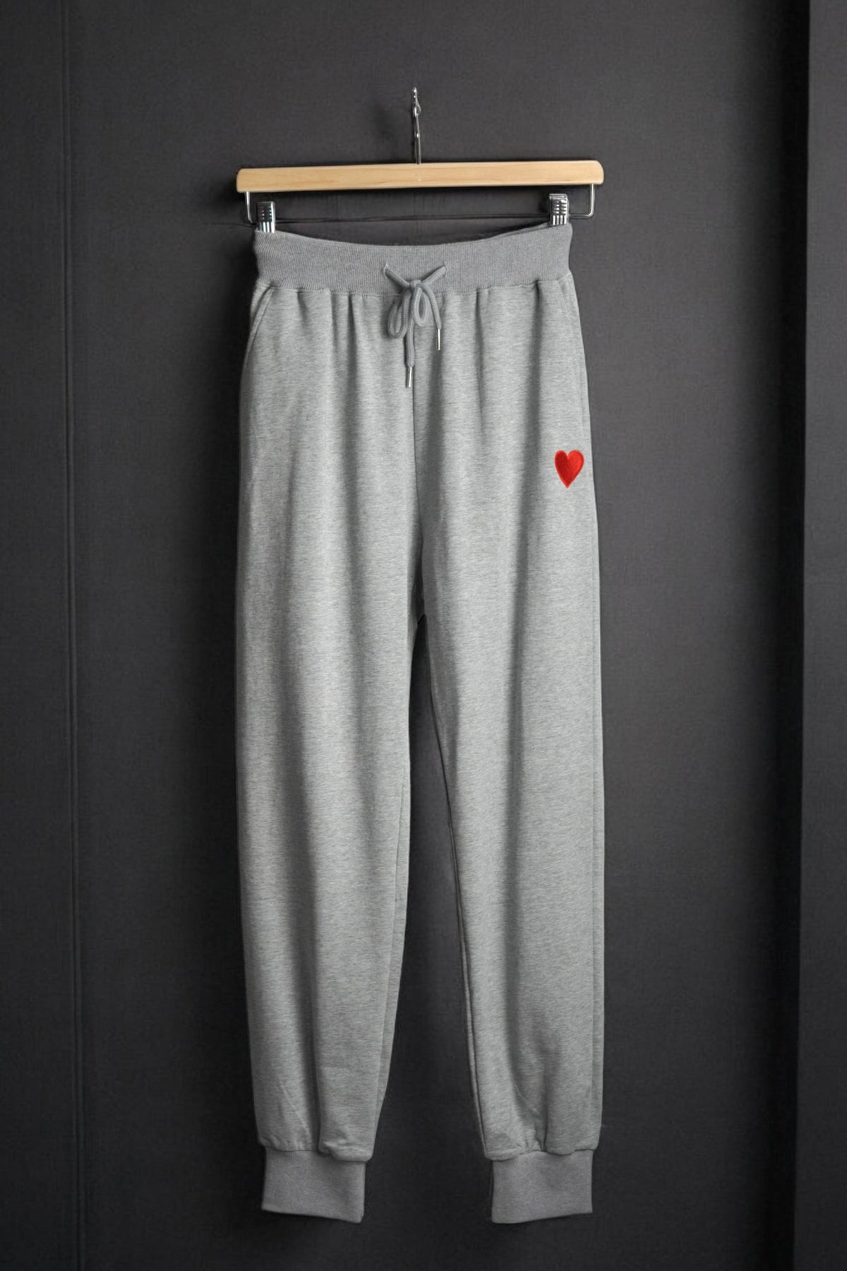 Pantaloni de trening gri cu talie elastică și șnur, decorați cu o inimă roșie brodată pe piciorul stâng.