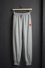 Pantaloni de trening gri cu talie elastică și șnur, decorați cu o inimă roșie brodată pe piciorul stâng.