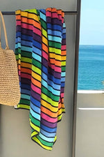 Prosop mare de plajă agățat pe un suport, cu dungi orizontale multicolore în nuanțe vibrante de albastru, verde, galben, roz și portocaliu, decorând un balcon cu vedere la mare.