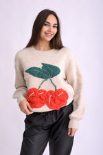 Detaliu de material tricotat bej cu aplicații tricotate în formă de cireșe și perle decorative.