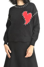 Vedere laterală a puloverului negru cu inimă roșie și cusături decorative albe pe umăr.