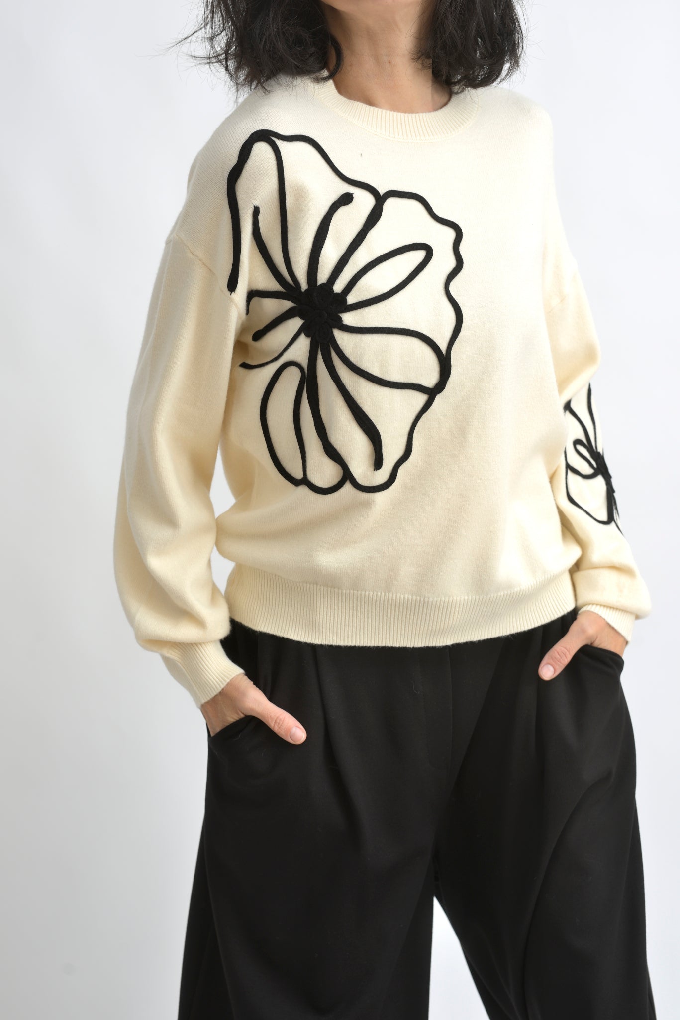 Pulover crem din tricot, cu aplicație florală mare din șnur negru pe față, purtat cu pantaloni negri.