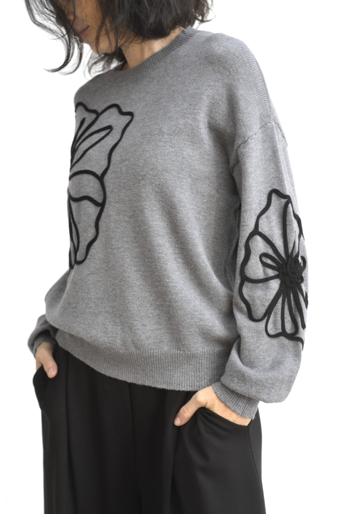 Pulover lejer din tricot gri, cu umeri căzuți și aplicație mare din șnur negru în formă de floare pe față.