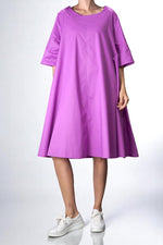Rochie oversized din bumbac într-o nuanță vibrantă de cyclamen, cu croi în A, buzunare laterale și mâneci trei sferturi. Ținută relaxată, purtată cu adidași albi.