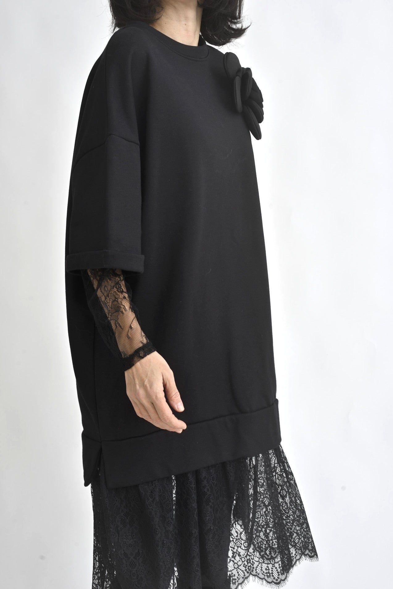 Rochie oversize neagră cu floare 3D aplicată pe umăr și finisaj din dantelă la tiv, surprinsă în unghi 3/4.