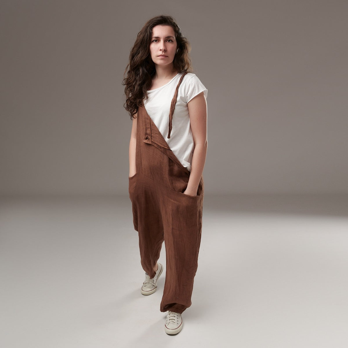 Salopetă lungă din in, culoare maro, cu bretele reglabile și croială lejeră, purtată cu tricou alb și pantofi sport albi