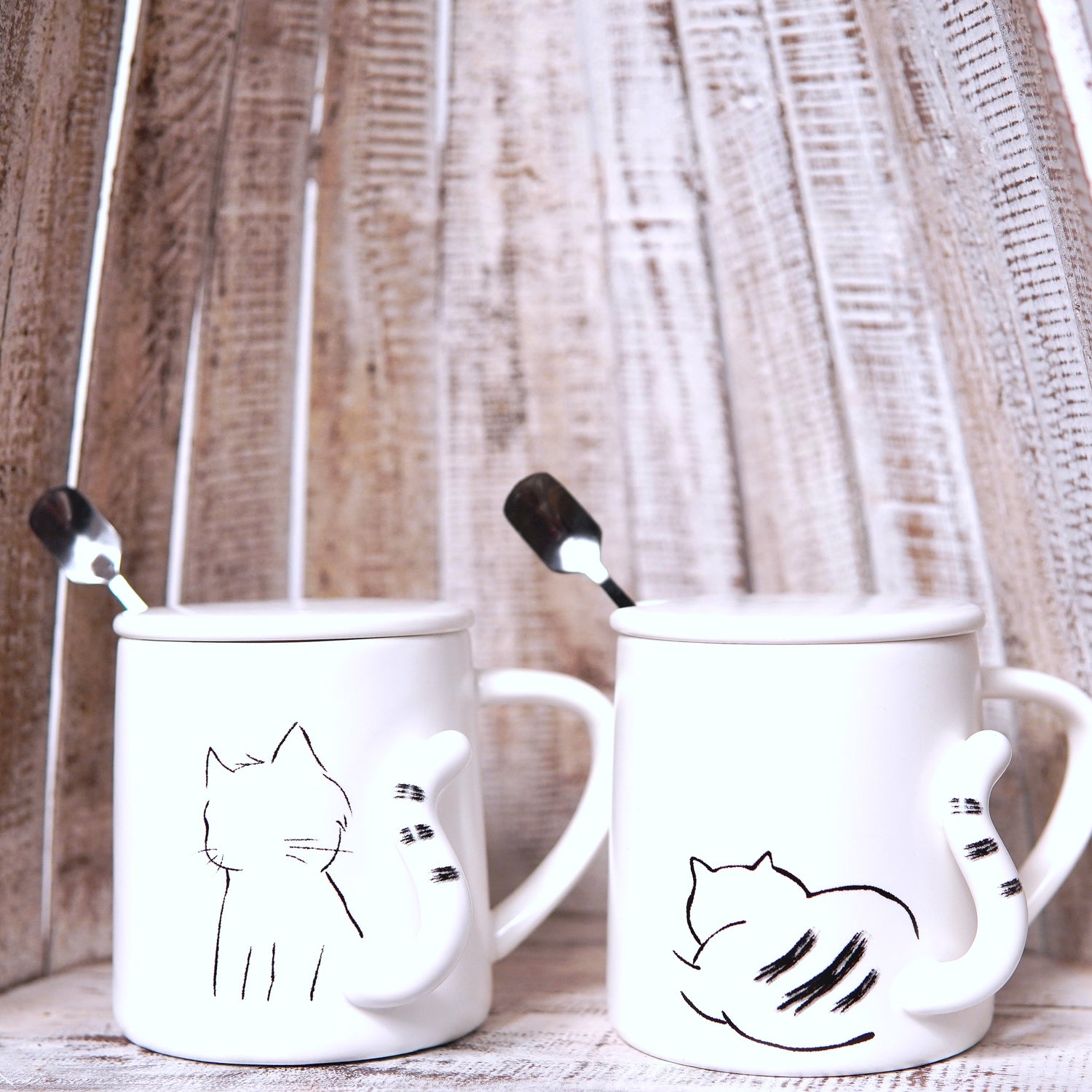 Set de două căni albe din ceramică, cu desene minimaliste de pisici și mânere în formă de coadă, cu lingurițe din inox.