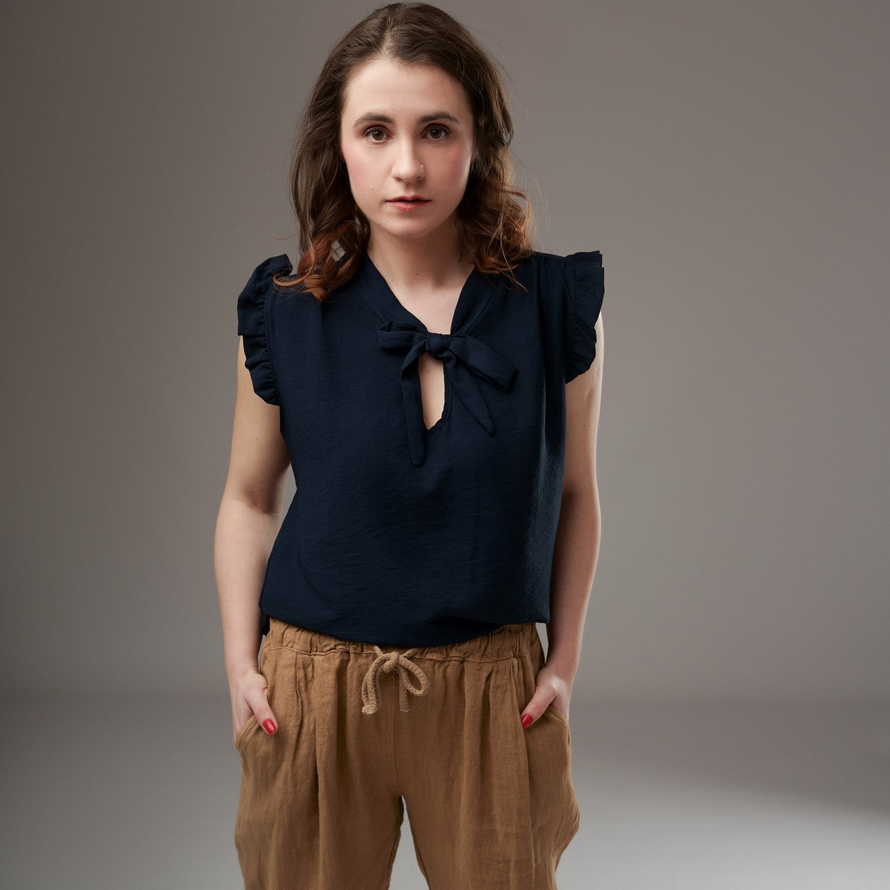 Top bleumarin fără mâneci, cu cravată la gât și volănașe, purtat de model cu pantaloni lejeri bej.