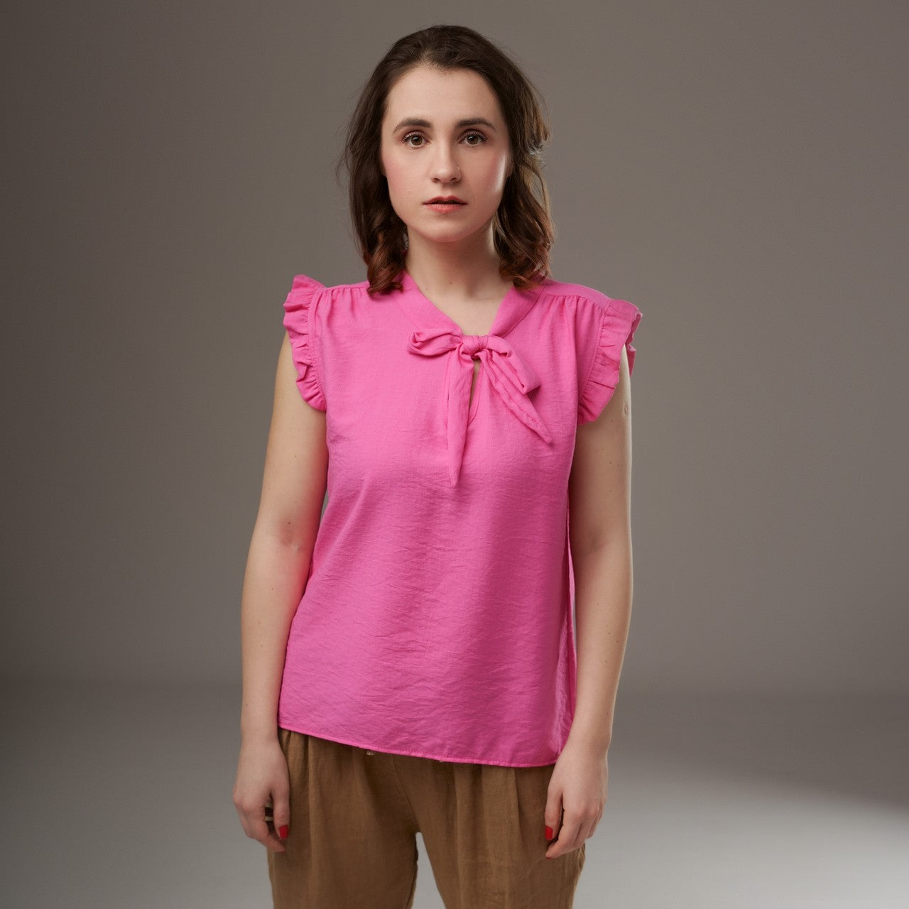 Top roz intens din viscoză, fără mâneci, cu cravată și volănașe, purtat cu pantaloni lejeri bej.