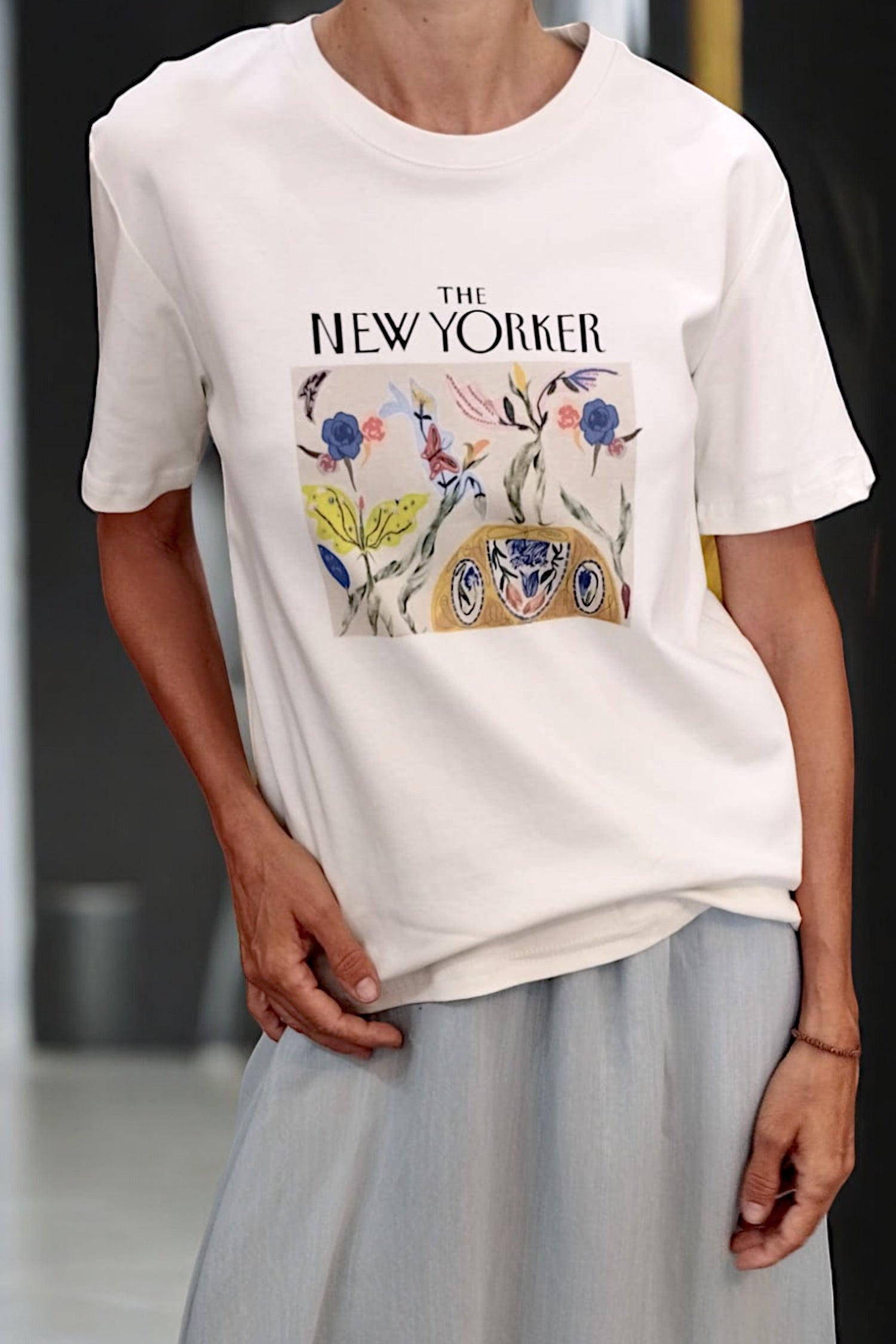 Detaliu al imprimeului „The New Yorker” cu fluturi, frunze și elemente florale colorate pe tricou alb.