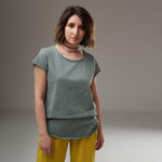 Tricou verde salvie cu decolteu rotund și margini rulate, purtat lejer cu pantaloni galbeni.