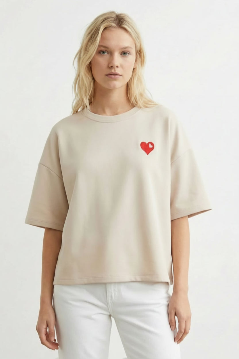 Tricou oversize bej cu inimioară roșie aplicată pe piept, purtat cu pantaloni albi, vedere frontală.