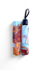 Umbrelă pliată cu imprimeu artistic multicolor, prevăzută cu husă asortată.