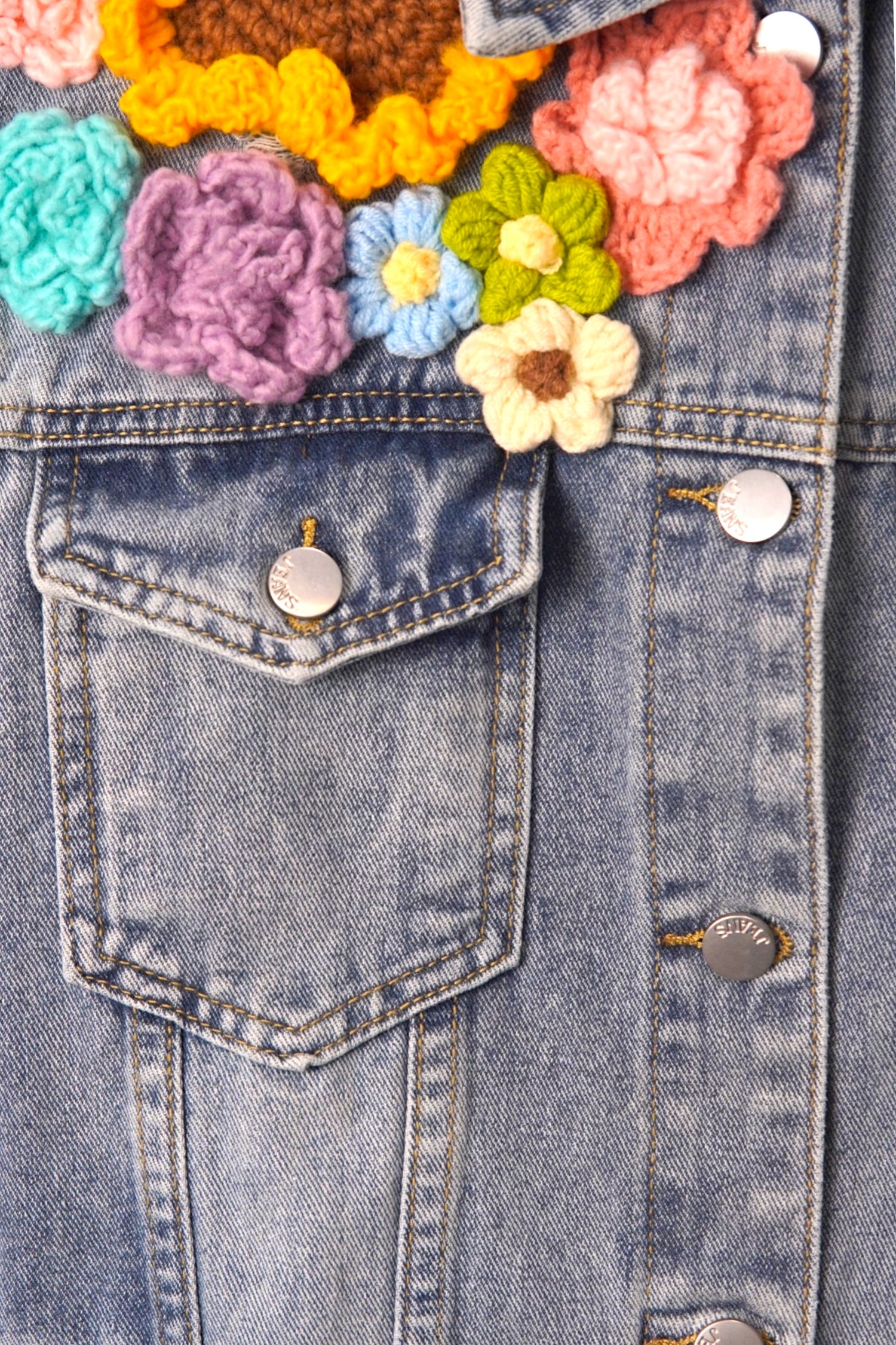 Buzunar frontal al unei veste din denim, decorat cu flori croșetate multicolore și nasturi metalici.