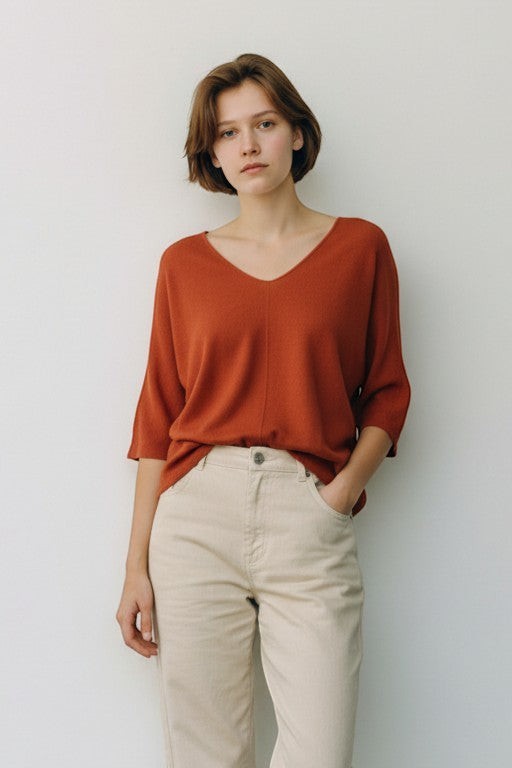 All Above Boutique rust orange relaxed fit V-neck sweater with dropped shoulders and 3/4 sleeves, paired with light beige trousers.