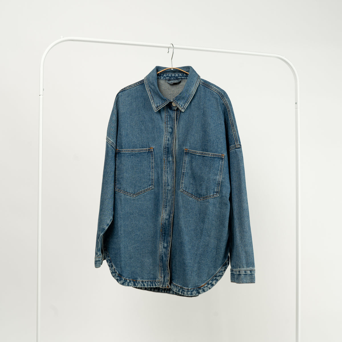  Jachetă tip cămașă din denim albastru, cu buzunare mari și închidere cu capse, expusă pe un umeraș alb.