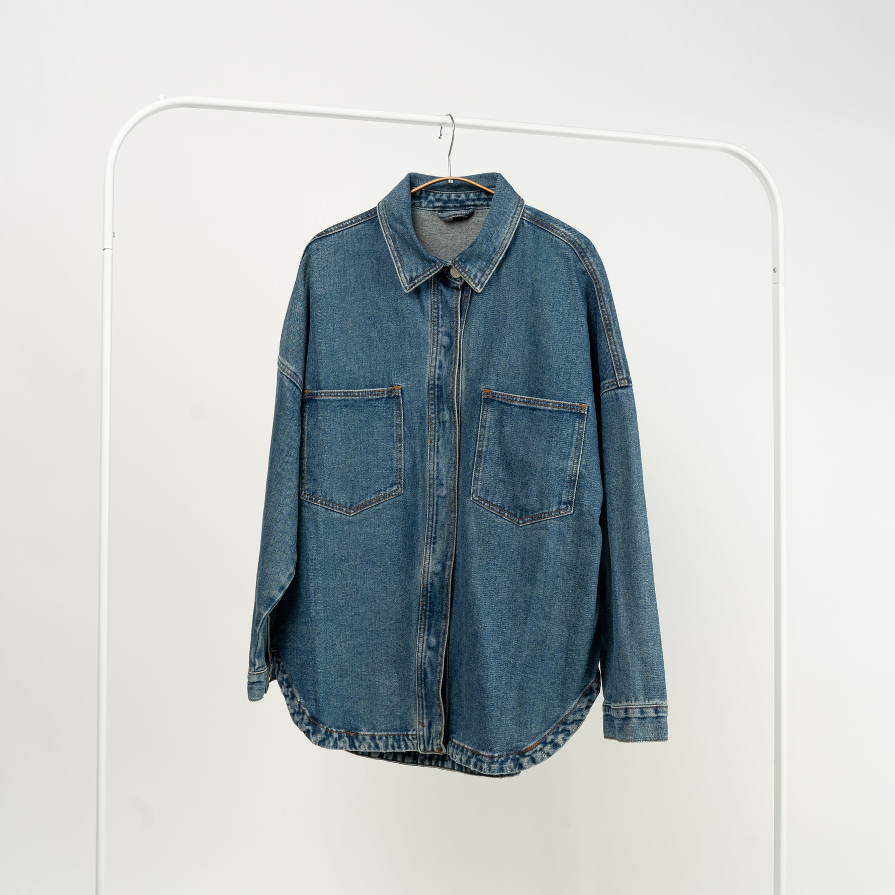  Jachetă tip cămașă din denim albastru, cu buzunare mari și închidere cu capse, expusă pe un umeraș alb.