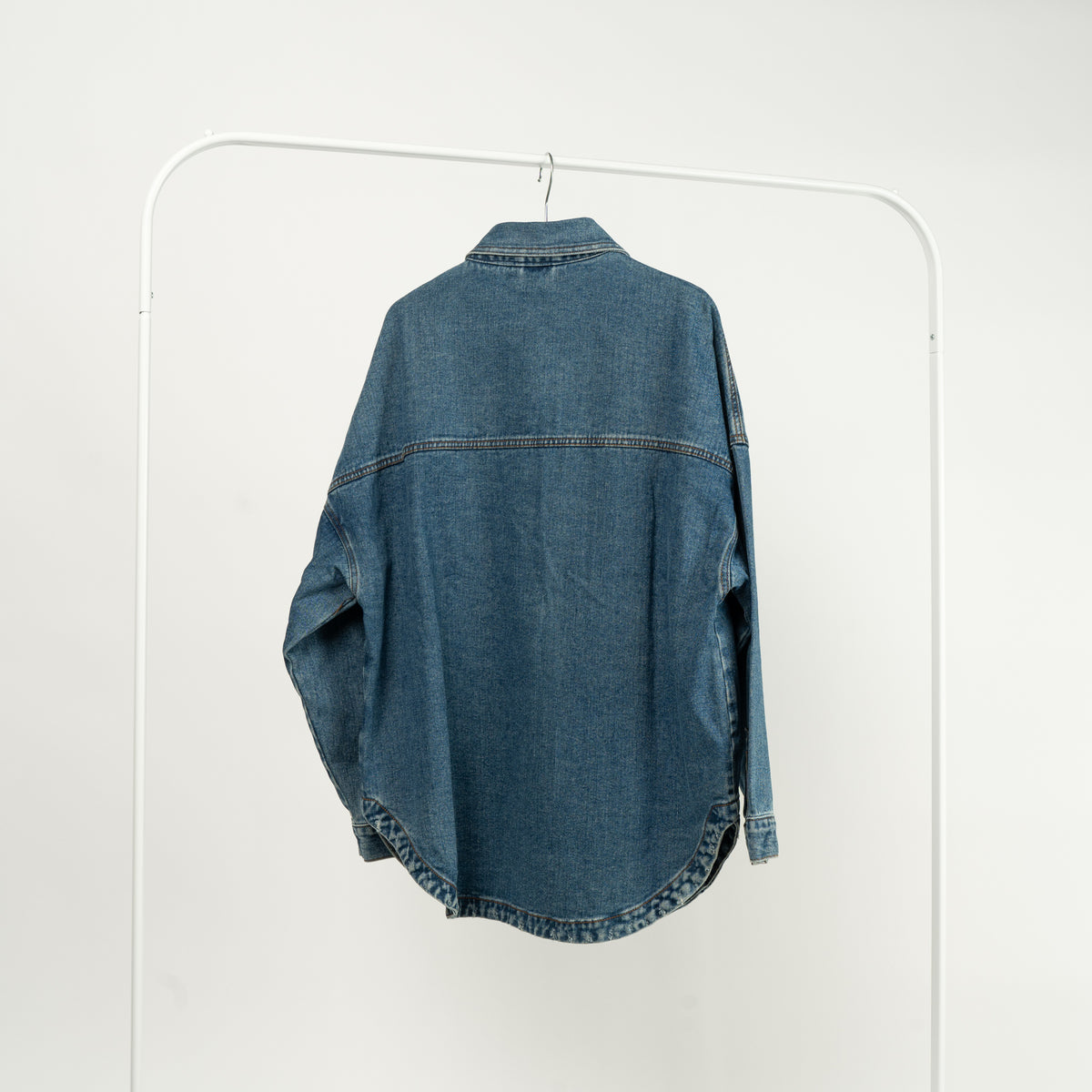  Jachetă tip cămașă din denim albastru, cu buzunare mari și închidere cu capse, expusă pe un umeraș alb, vedere spate