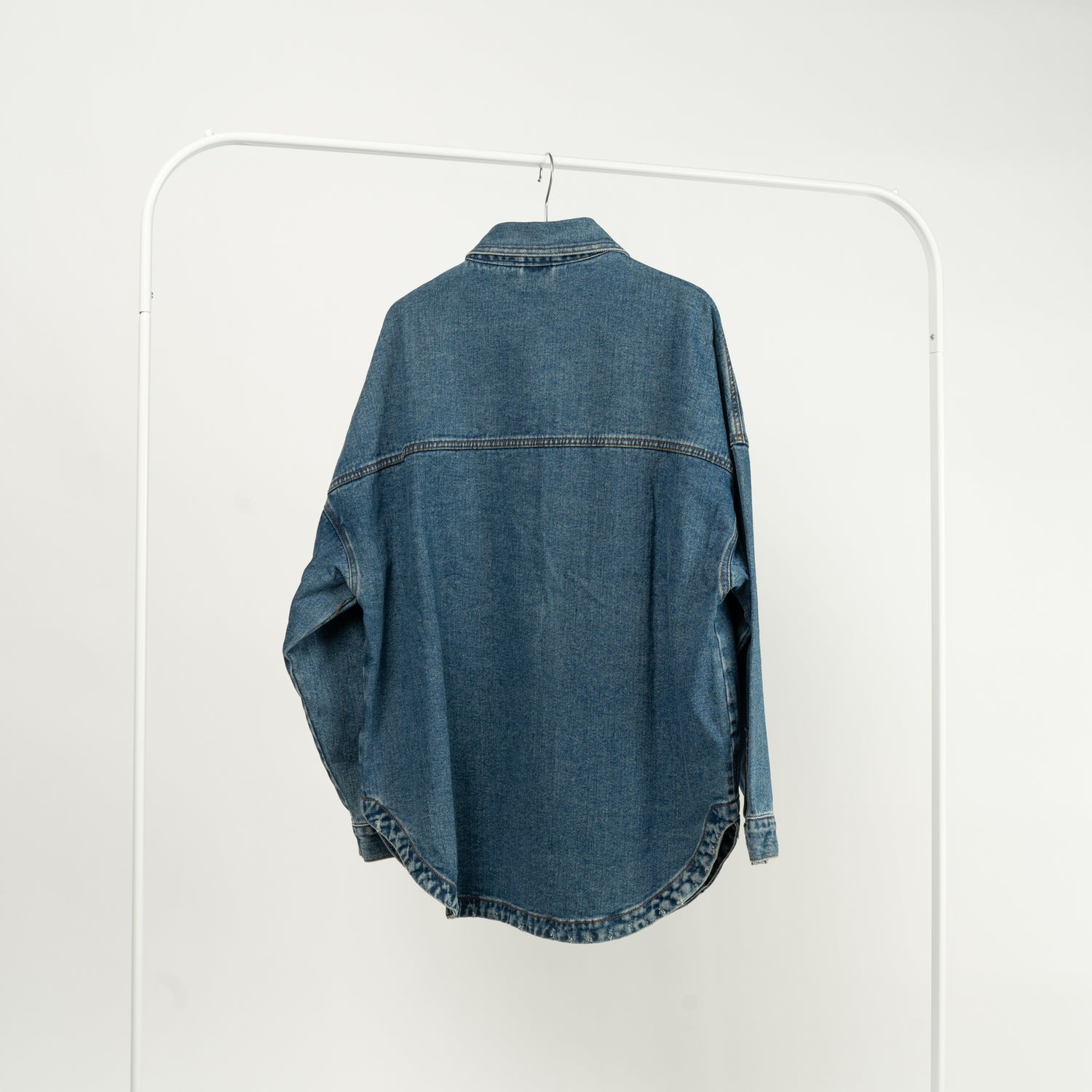 Jachetă tip cămașă din denim albastru, cu buzunare mari și închidere cu capse, expusă pe un umeraș alb, vedere spate