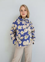 Model purtând jachetă damă cu imprimeu floral bej pe fundal albastru, croială asimetrică și guler înalt